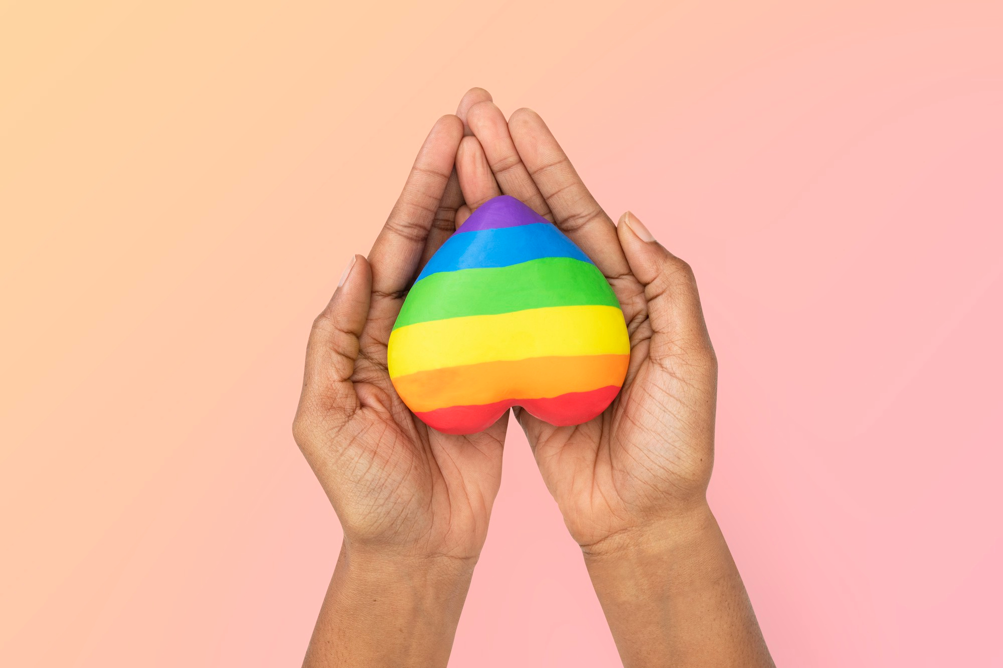 Imagem de duas mãos, em um fundo rosa, seguranfo um coração com as cores da bandeira LGBT, para simbolizar o Dia Internacional do Orgulho LGBTQIAP+